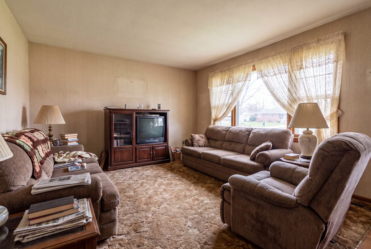 A dated living room with brown sofas, a recliner, an old TV cabinet, lace curtains and a shag carpet.